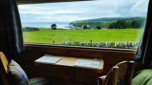 A cabin with a wooden desk and two plaid chairs, a window with a view of a green field and a stone wall, and a map on the desk. - Bespoke Hand Built Shepherds Hut in Dunbeath (Dunbeath)