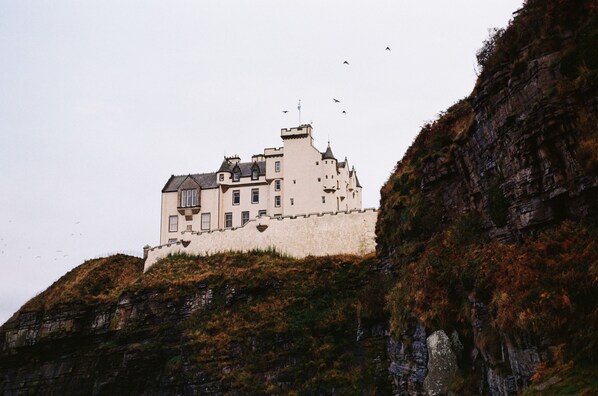 A castle-like building with multiple towers and a fortified wall, situated on a rocky hill.