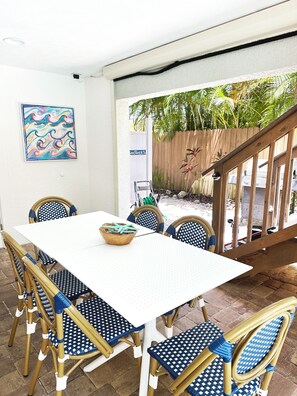 Un patio con una mesa blanca y sillas de mimbre a cuadros azules y blancos, una cesta tejida y una valla de madera con plantas.