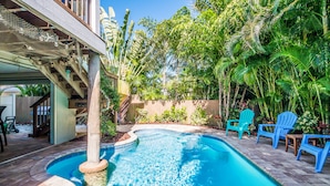 Ein Pool mit Holzdeck, umgeben von tropischen Pflanzen und blauen Stühlen.