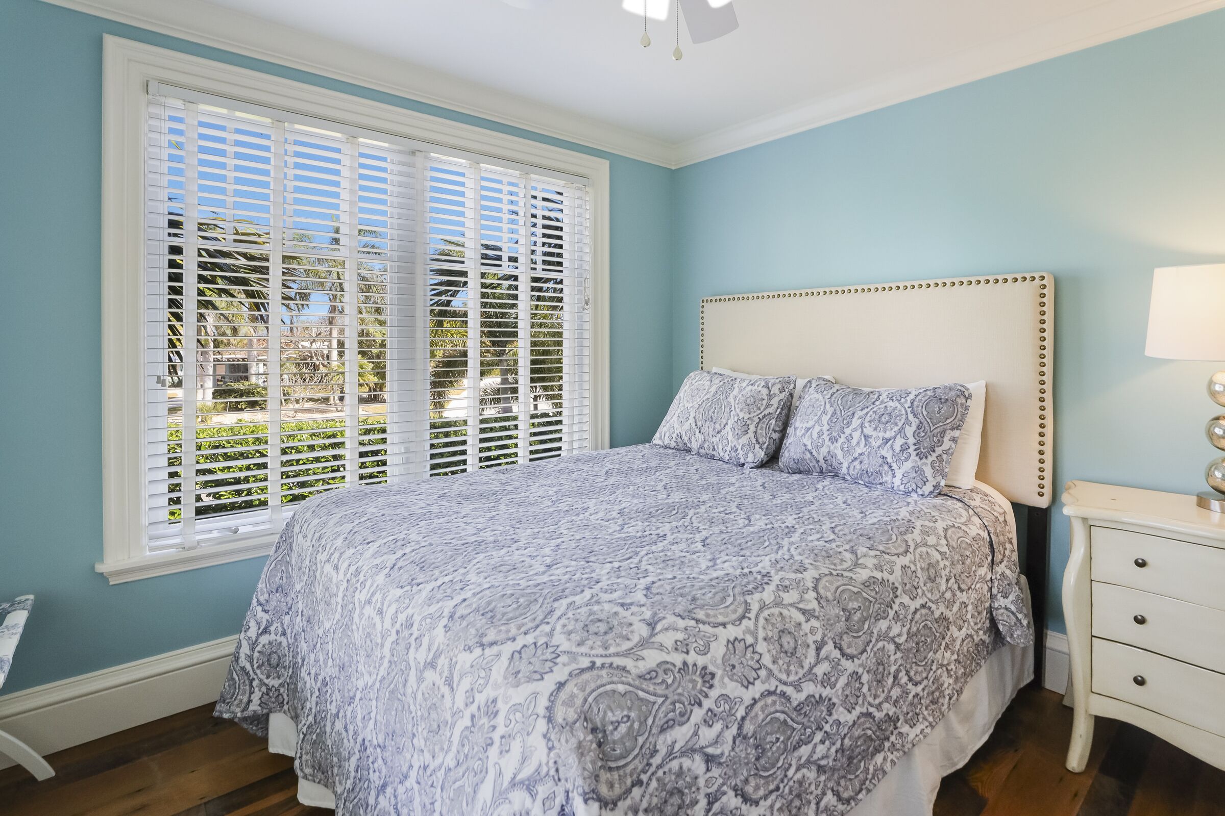 A bedroom with a bed, a nightstand, a window with blinds, and a view of greenery outside.