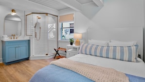 Een slaapkamer met een bed, een nachtkastje met een lamp, een raam met uitzicht, een douche en een wastafel met spiegel.