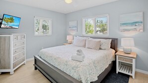 Un dormitorio con cama, televisor, dos ventanas con persianas, ventilador de techo y mesita de noche con lámpara.