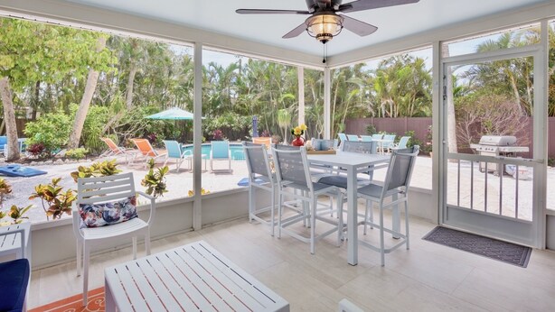 Un patio techado con mesa y sillas, un ventilador de techo, y vistas a una piscina y jardín.