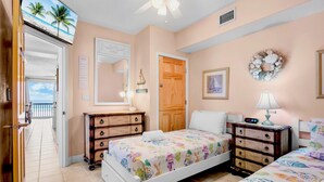 A bedroom with two beds, a wooden dresser, a nightstand, a wall clock, and a view of the ocean through an open door. - Beachfront Rental with Ocean Views, Close to Restaurants and Shopping! (Bradenton Beach)