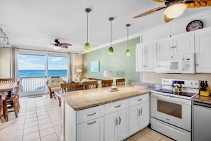 A kitchen with a large island, white cabinets, a microwave, and a stove. There is a dining area with a table and chairs, and a view of the ocean through the sliding glass doors.