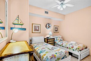 A bedroom with two beds, a nightstand, a lamp, a ceiling fan, and wall decorations including a sailboat and framed pictures.
