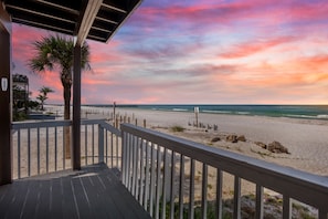 A beach view from a wooden deck with a railing, palm trees, and a sunset sky.