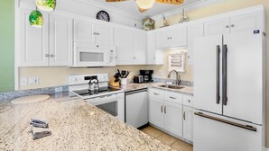 A kitchen with white cabinets, a stainless steel refrigerator, a microwave, a stove, and a ceiling fan with leaf designs. - Beachfront Rental with Ocean Views, Close to Restaurants and Shopping! (Bradenton Beach)