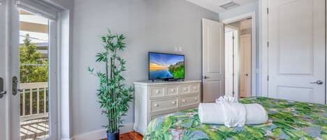 A bedroom with a bed, a television on a dresser, a potted plant, and a door leading to another room.