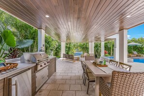 Cocina exterior con asador, zona de comedor y vista a la piscina.