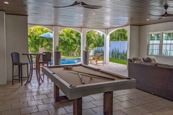 A pool table with surrounding seating, a ceiling fan, and a view of a swimming pool and greenery.