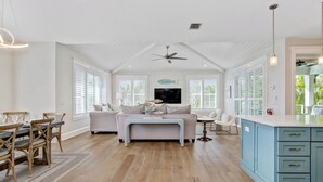 A spacious living room with a high ceiling, a ceiling fan, and a large window with blinds. There is a dining area with wooden chairs and a table, and a kitchen area with blue cabinets.