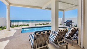 Zona junto a la piscina con sillones de descanso, una parrilla y vistas a la playa.