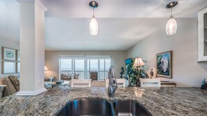 A kitchen with granite countertops, stainless steel sink, pendant lighting, and a view to the living area.