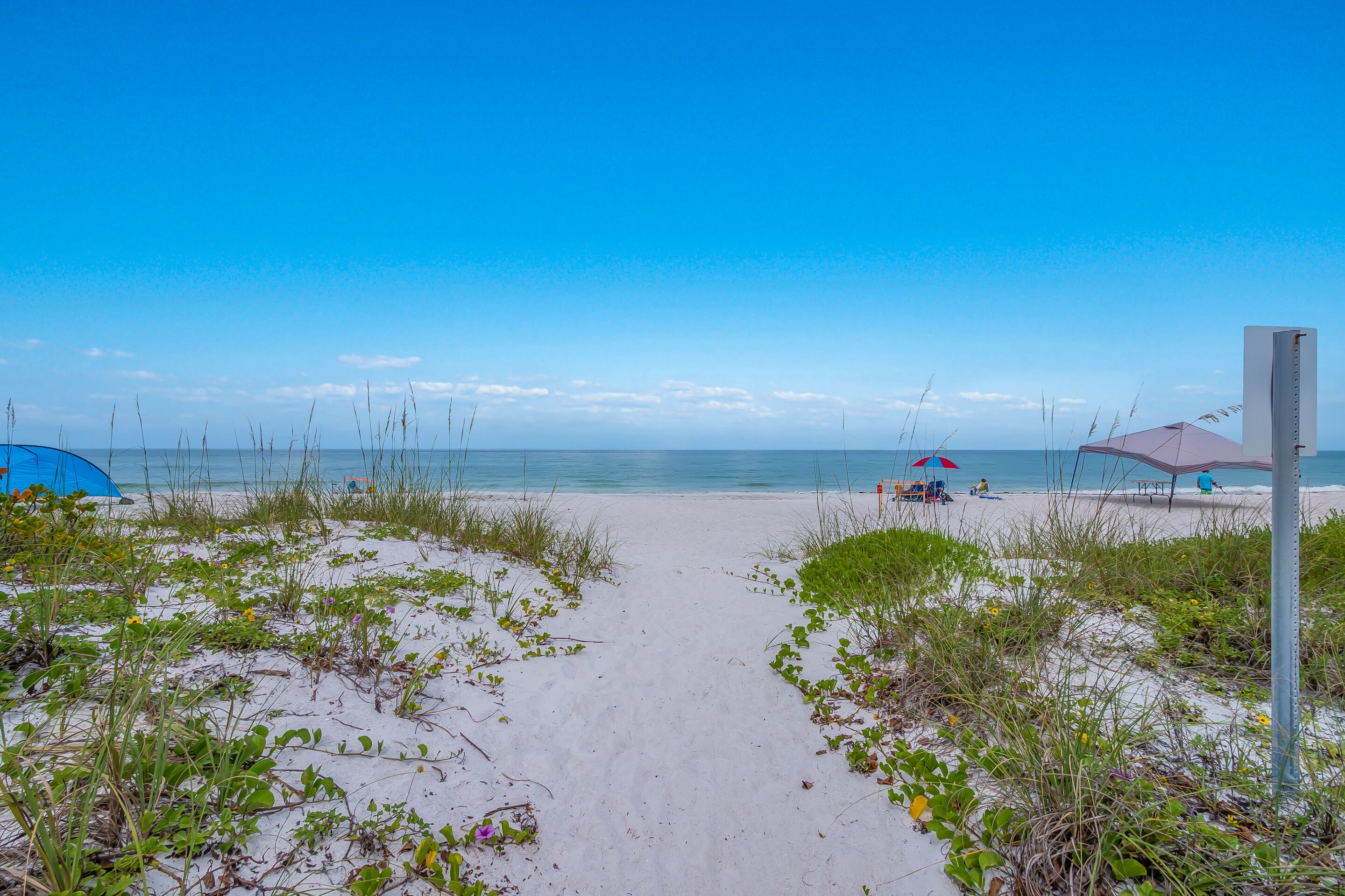 Un sentier qui longe la plage et mène à l’océan, avec une tente et des parasols en arrière-plan.