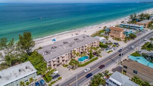 An aerial view of a coastal area with a beach, a swimming pool, and a multi-story building with parking.