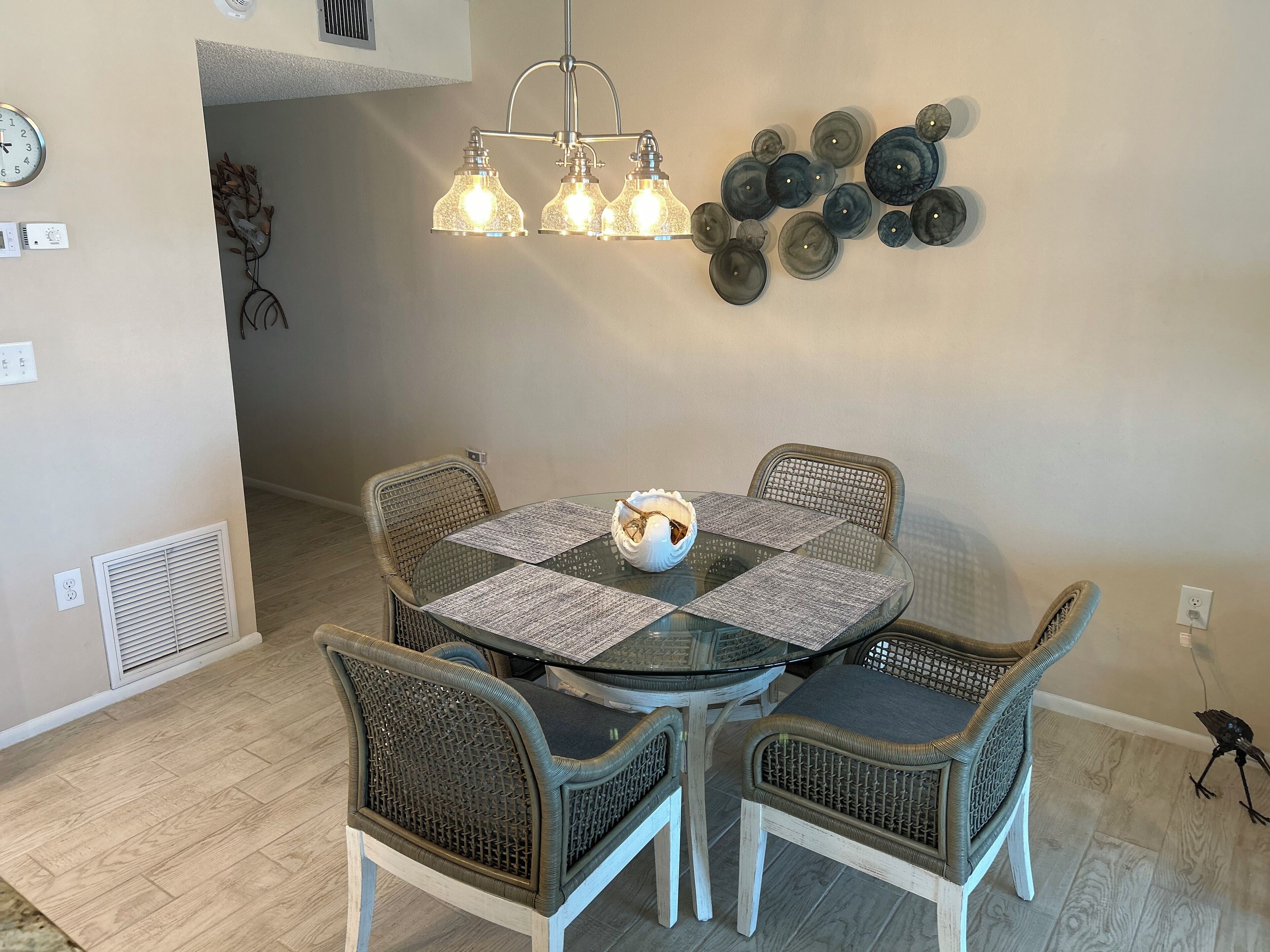 Un coin repas comprenant une table ronde en verre, quatre chaises en osier, une œuvre d’art décorative accrochée au mur et un luminaire suspendu.
