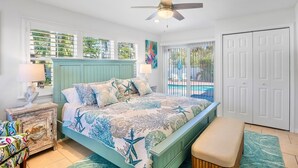 Un dormitorio con cama, una banca, una mesita de noche, ventilador de techo y vista a una piscina.