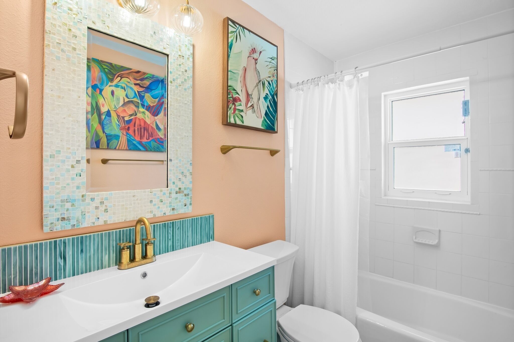 A bathroom with a white sink, teal vanity, and a white toilet. There are two framed paintings on the wall and a window with curtains.