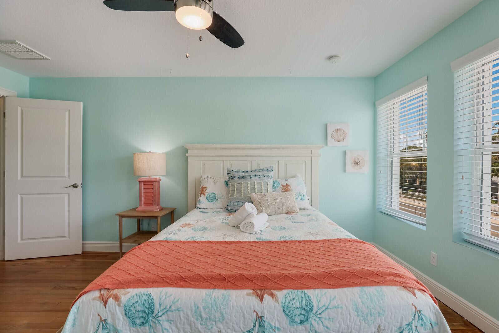 A bedroom with a bed, a ceiling fan, a nightstand with a lamp, and a window with blinds.