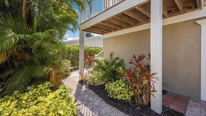 A covered outdoor area with a pathway, surrounded by various tropical plants and a white building structure. - Coastal Home with Heated Pool, Close to Beach! (Holmes Beach)