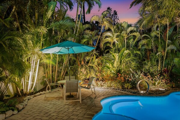 Zona de piscina con mesa y sillas, rodeada de exuberante vegetación tropical.