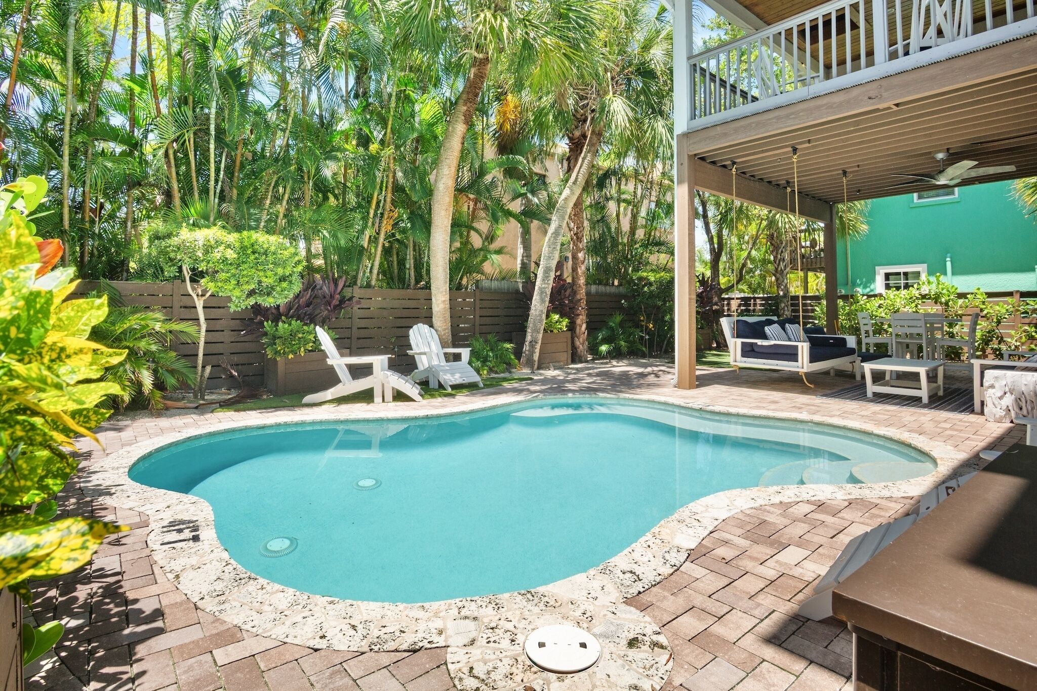A swimming pool with a brick deck, surrounded by palm trees and a wooden deck with outdoor furniture.