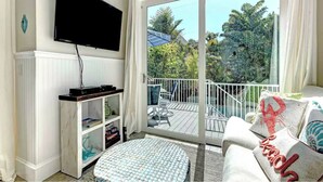 A living room with a flat-screen TV mounted on the wall, a white entertainment unit with shelves, a round ottoman, and a balcony with a white railing and outdoor furniture. - Villa with Heated Pool and Swim-up Tiki Bar! Close to Beach - La Mer (Holmes Beach)