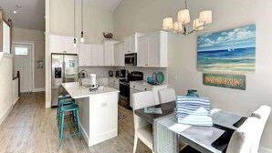 A modern kitchen with white cabinets, a central island with bar stools, a dining area with a table and chairs, and a painting of a beach scene. - Villa with Heated Pool and Swim-up Tiki Bar! Close to Beach - La Mer (Holmes Beach)