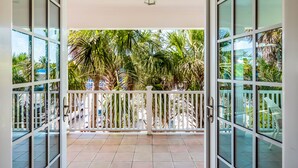 A balcony with glass doors, white railings, and a view of palm trees and parked cars. - Cozy Pine Ave Cottage Near Beach (Anna Maria)