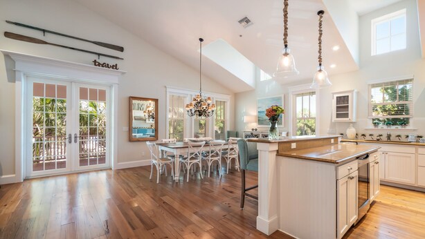 A spacious kitchen with wooden flooring, a central island, pendant lighting, and large windows offering views of greenery. - Cozy Pine Ave Cottage Near Beach (Anna Maria)