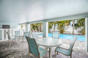 Un patio exterior con piscina, zona de estar cubierta y vistas a palmeras y una cerca blanca.