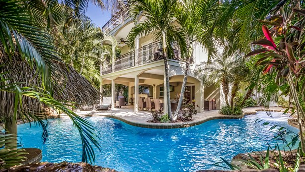A pool area with a curved design, surrounded by palm trees and a multi-level building with outdoor seating. - It's a Jungle Out There - Tropical Pool, Sunset Views, Game Room! (Holmes Beach)