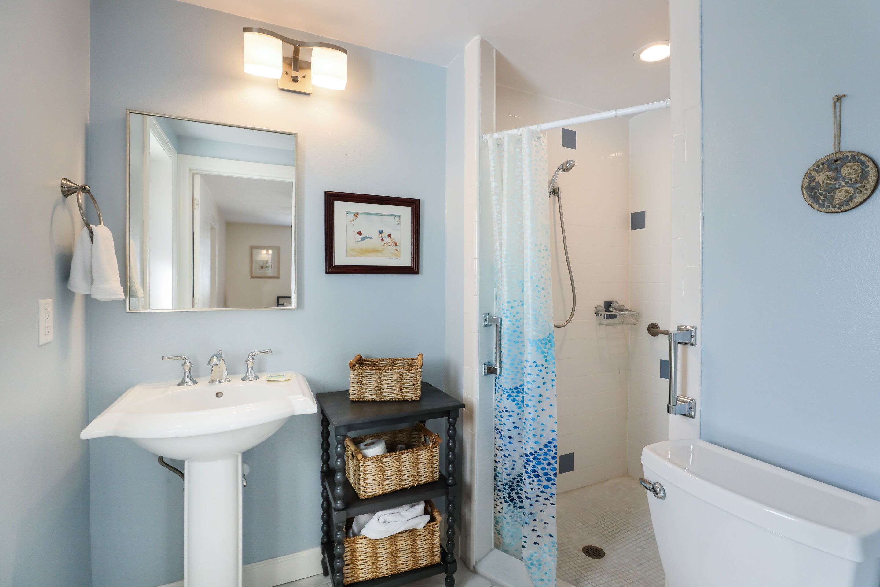 A bathroom with a white sink, a mirror, a framed picture, a black vanity with wicker baskets, and a shower with a patterned curtain.