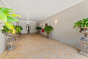 A hallway with tiled flooring, potted plants, a statue, and wall-mounted lights.