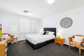A bedroom with a bed, bedside tables, a desk, wicker chairs, a wall art piece, and a window with blinds.