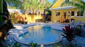 A pool area with a yellow house, lounge chairs, and tropical plants. - Covey Cottage! Walk to Beach, Boutique Shops and City Pier (Anna Maria)