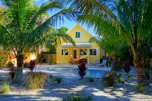 Une maison jaune avec une piscine, entourée de palmiers et de différentes plantes.