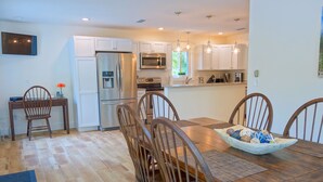 A modern kitchen with white cabinets, stainless steel appliances, and a dining area with wooden chairs and a large table. - Covey Cottage! Walk to Beach, Boutique Shops and City Pier (Anna Maria)