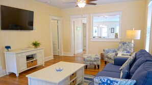 A living room with a TV, white furniture, a ceiling fan, and a large window. - Covey Cottage! Walk to Beach, Boutique Shops and City Pier (Anna Maria)