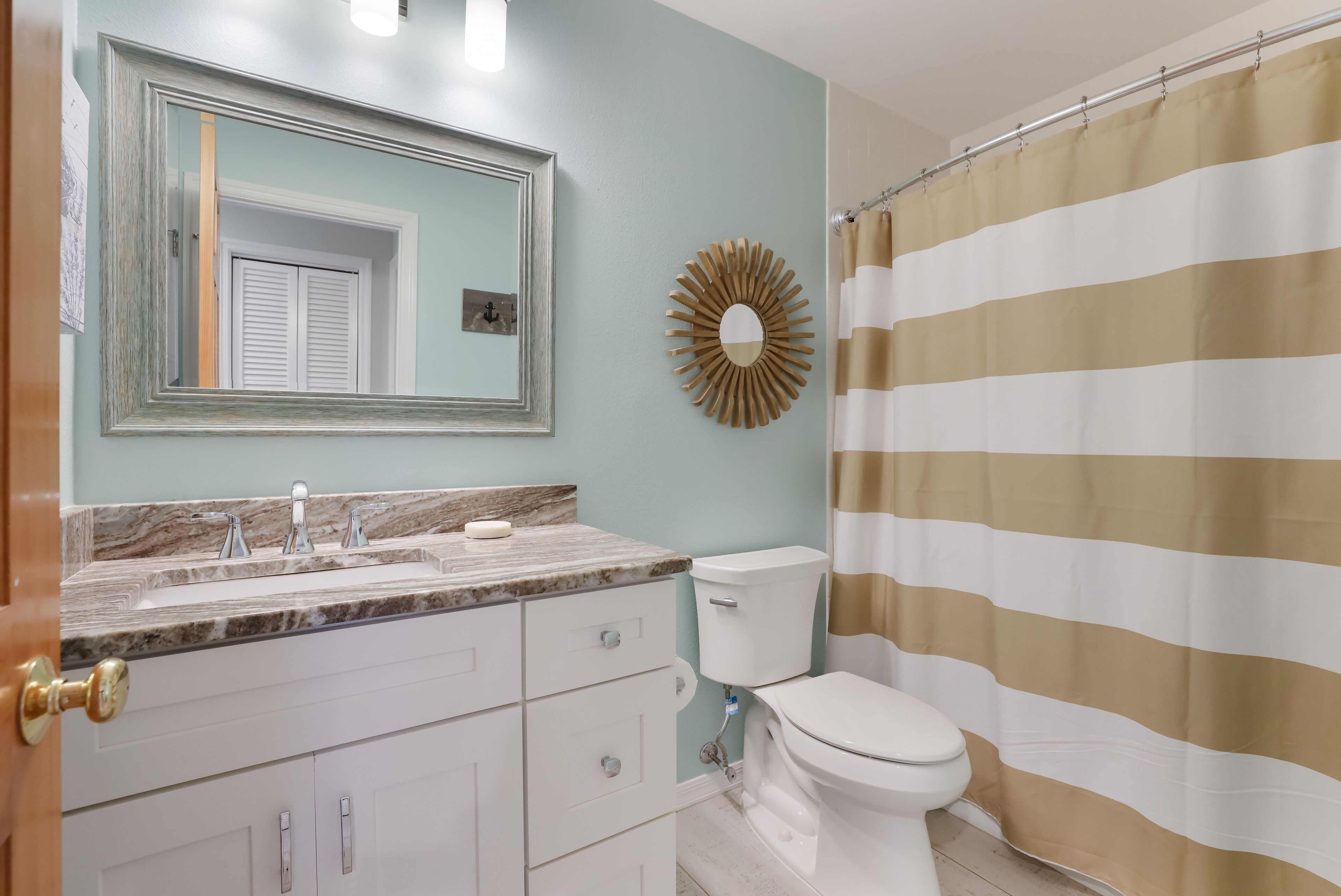 A bathroom with a marble sink, a toilet, a shower curtain, and a mirror with a sunburst decoration.