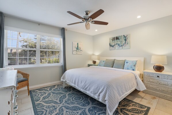 A bedroom with a bed, a ceiling fan, a window with blinds, a desk, and a patterned rug.