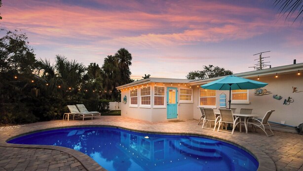 A backyard with a swimming pool, a patio with chairs, and a house with a blue door.