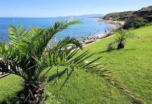 A coastal landscape with a grassy area, palm trees, and a beach with people. - Mediterranean Vacation Homes by the Sea in a Quiet Location, with a Garden & BBQ (Sardinien)