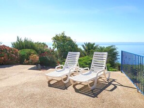 Zwei weiße Lounge-Stühle mit Metallgestellen auf einem Kiesweg, mit Blick auf Meer und grüne Landschaft im Hintergrund.