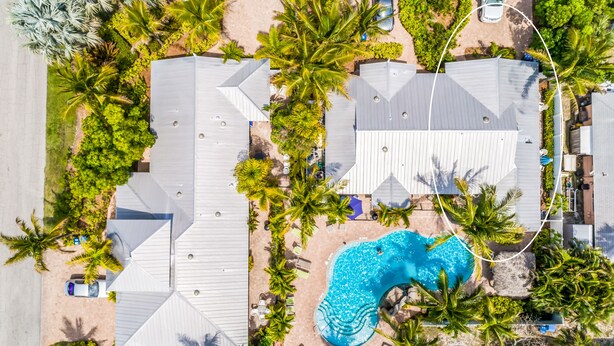 Vue aérienne d’un quartier résidentiel avec une piscine entourée de palmiers et de maisons aux toits inclinés.