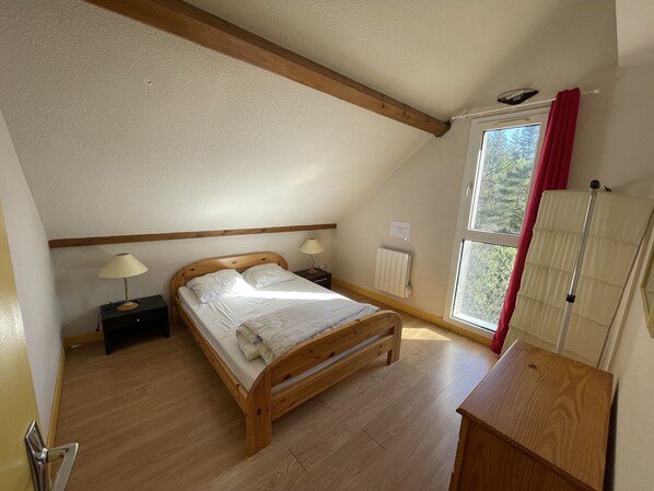 A bedroom with a wooden bed, two bedside lamps, a radiator, a window with a view of trees, and a wooden dresser. - Appartement T3 résidence porte du Sud (Le Lioran)
