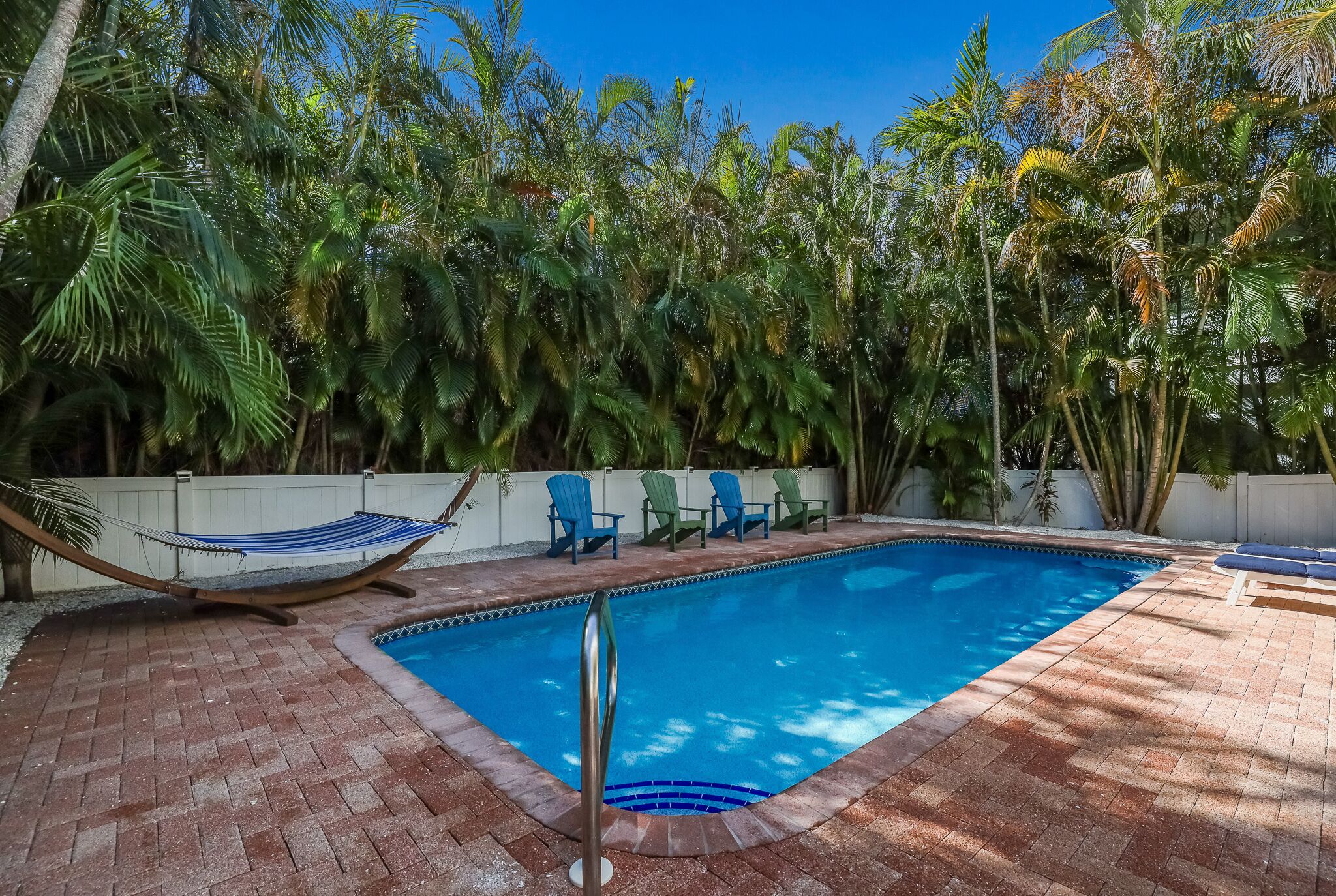 A rectangular swimming pool with a brick border, surrounded by palm trees and a hammock.