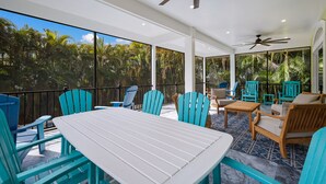 Une vaste terrasse extérieure dotée d’une table blanche et de chaises bleues, entourée de grandes fenêtres qui offrent une vue sur une végétation luxuriante.